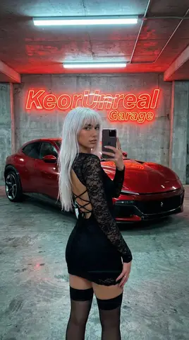 Женщина в чёрном платье делает селфи в гараже с красным Ferrari и вывеской KeorUnreal Garage.
