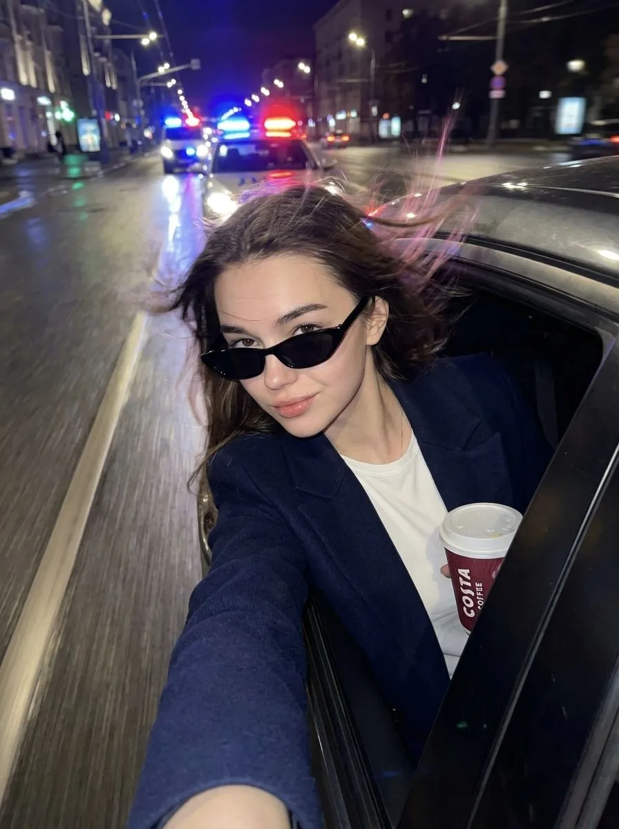 Женщина с телефоном высунулась из окна автомобиля на мокрой ночной улице с полицейскими огнями.