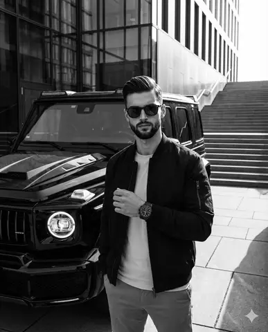 Мужчина в тёмной куртке и очках стоит рядом с чёрным Mercedes-Benz G-Wagon на залитой солнцем площади.