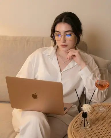 Женщина с золотистыми очками работает за MacBook Pro на светлом диване рядом с бокалом розового вина
