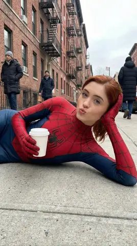 Рыжеволосая девушка в красно-синем костюме Spider-Man стоит на улице Нью-Йорка с кофе и показывает знак мира.