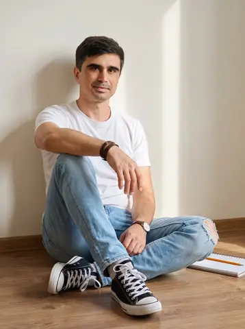 Человек в белой футболке, светлых джинсах и кедах Converse сидит у светлой стены.