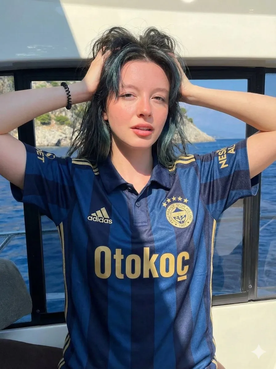 Молодая женщина в футболке Fenerbahçe позирует на яхте у окна с видом на море.