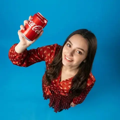 Улыбающаяся женщина поднимает красную банку Coca-Cola к камере на синем фоне.