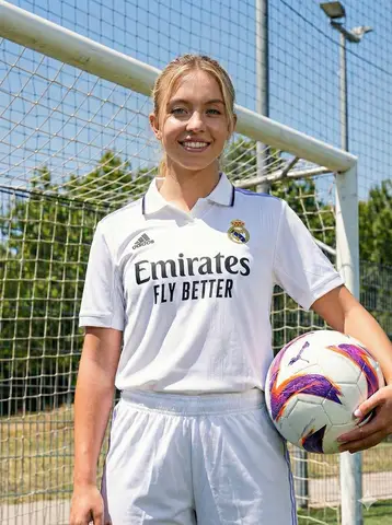 Девушка в белой форме Real Madrid держит футбольный мяч на солнечном тренировочном поле.