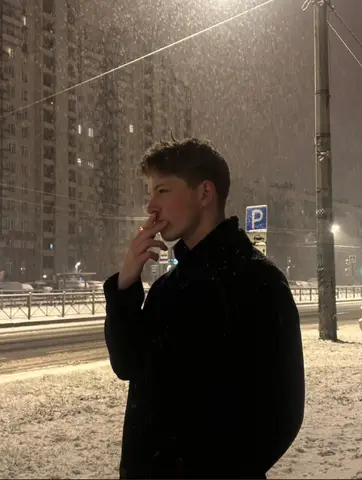 Молодой мужчина в тёмном пальто стоит в профиль, держа сигарету у рта на фоне ночного снегопада.