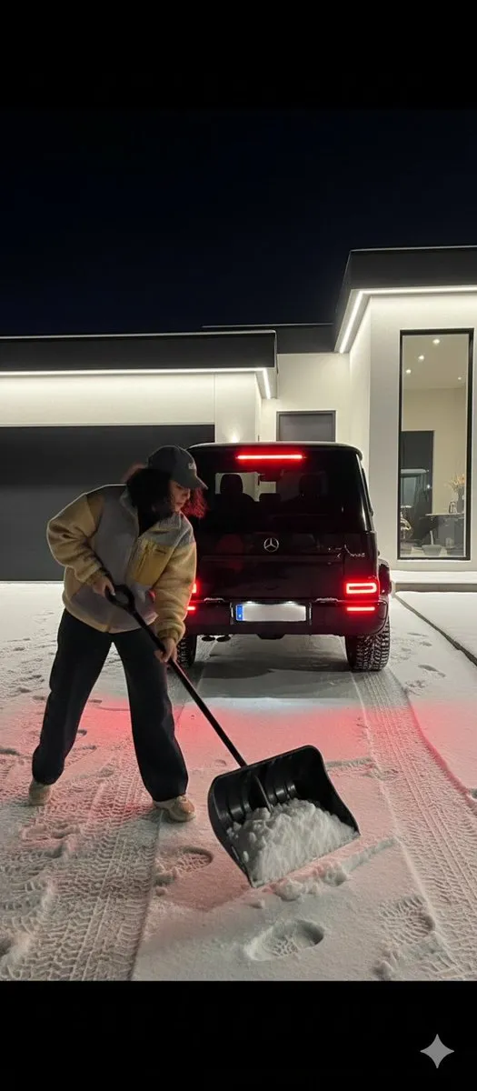 Женщина убирает снег перед чёрным Mercedes-Benz G 63 AMG у современного дома.
