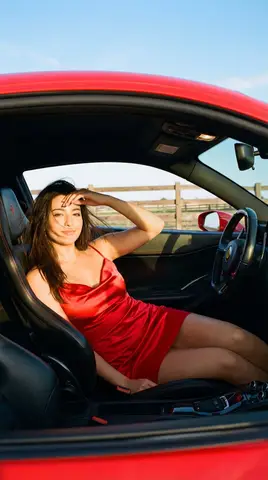 Женщина в красном атласном платье отдыхает в салоне Ferrari при тёплом солнечном свете.