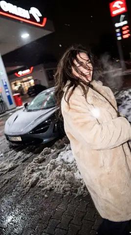 Женщина в объёмном меховом пальто смеётся у McLaren на заснеженной заправке ORLEN ночью.