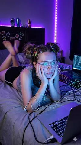 22-летняя женщина в gamer-girl образе лежит среди светящихся экранов в синей и пурпурной подсветке.