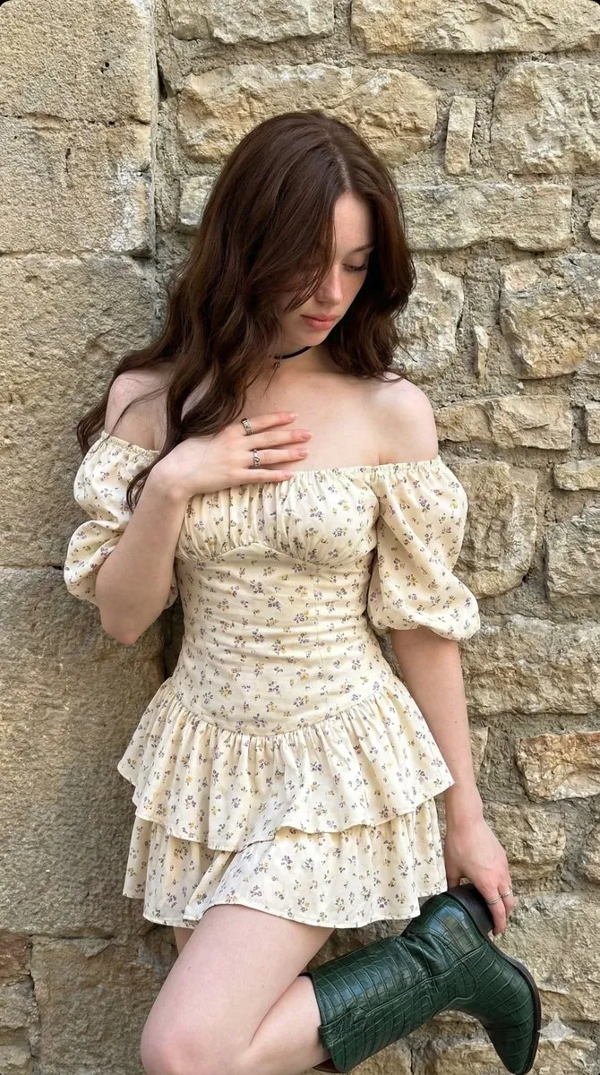 Женщина в кремовом платье держит тёмно-зелёный ковбойский сапог у каменной стены.