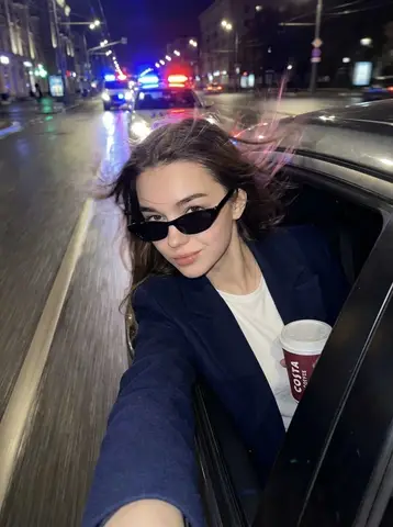 Женщина с телефоном высунулась из окна автомобиля на мокрой ночной улице с полицейскими огнями.